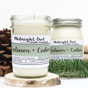 K&ouml;nnte beinhalten: Zwei Glasbeh&auml;lter mit wei&szlig;en Kerzen darin. Die Beh&auml;lter sind mit "Midnight Owl Candle Company" und "Balsam + Cedar" beschriftet. Die Beh&auml;lter stehen auf einer Holzoberfl&auml;che mit Kiefern&auml;sten.