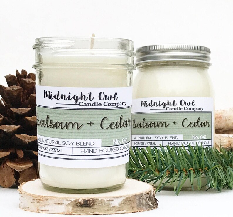 K&ouml;nnte beinhalten: Zwei Glasbeh&auml;lter mit wei&szlig;en Kerzen darin. Die Beh&auml;lter sind mit "Midnight Owl Candle Company" und "Balsam + Cedar" beschriftet. Die Beh&auml;lter stehen auf einer Holzoberfl&auml;che mit Kiefern&auml;sten.