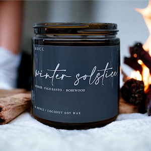 May include: A dark brown glass jar candle with a black label that reads "winter solstice" and "vetiver - palo santo - rosewood". The label also says "8 ounce / coconut soy wax".