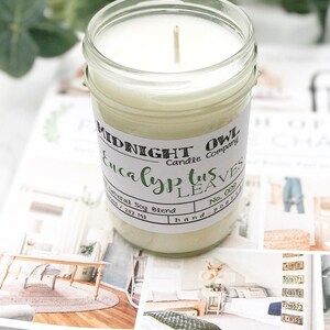 May include: A clear glass jar candle with a white wax and a wooden wick. The label reads "Midnight Owl Candle Company Eucalyptus Leaves No. 009 hand-poured".