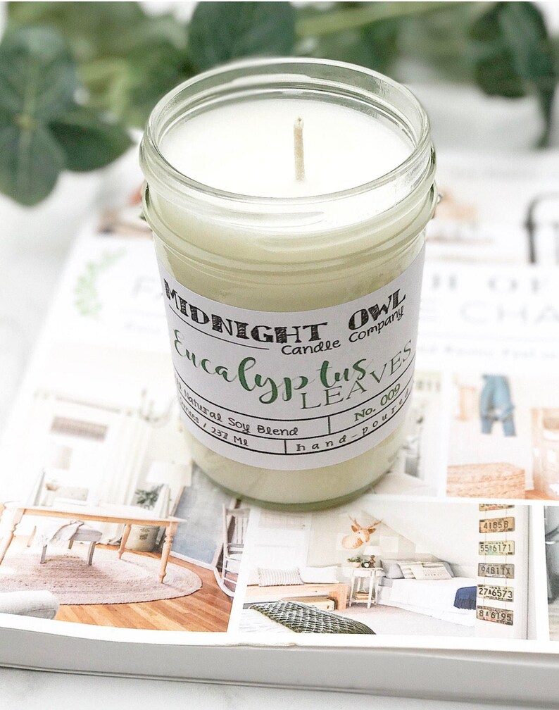 May include: A clear glass jar candle with a white wax and a wooden wick. The label reads "Midnight Owl Candle Company Eucalyptus Leaves No. 009 hand-poured".