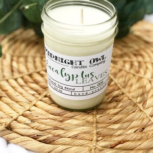May include: A white candle in a clear glass jar with a white label that reads "MIDNIGHT OWL Candle Company Eucalyptus Leaves No. 009 hand-poured". The candle is sitting on a woven straw mat.