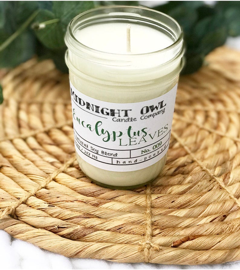 May include: A white candle in a clear glass jar with a white label that reads "MIDNIGHT OWL Candle Company Eucalyptus Leaves No. 009 hand-poured". The candle is sitting on a woven straw mat.