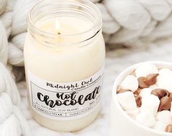 Könnte beinhalten: Eine brennende Sojakerze in einem Glas mit der Aufschrift "Midnight Owl Candle Company Chocolate". Die Kerze steht neben einer Tasse heißer Schokolade mit Marshmallows und Schokoladenstückchen. Der Hintergrund ist eine weiße, strukturierte Oberfläche.