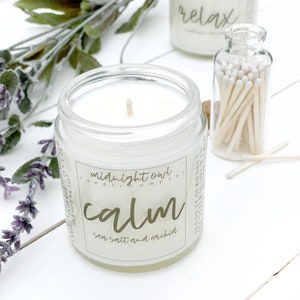 May include: A clear glass jar candle with a white wax interior and a gold label that reads "midnight owl candle company calm sea salt and orchid".