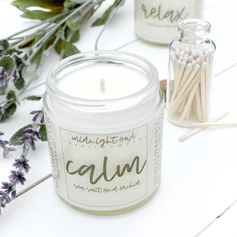 May include: A clear glass jar candle with a white wax interior and a gold label that reads "midnight owl candle company calm sea salt and orchid".