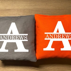 May include: Two square bean bags, one gray and one orange, are displayed. Each bag features a large white letter "A" with the name "ANDREWS" printed below. The bags are likely for a game like cornhole.