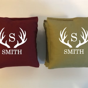 May include: Two square throw pillows, one burgundy and one olive green. Each pillow features a white antler design with the letter "S" in the center and the name "SMITH" below. The pillows are likely for home decor.