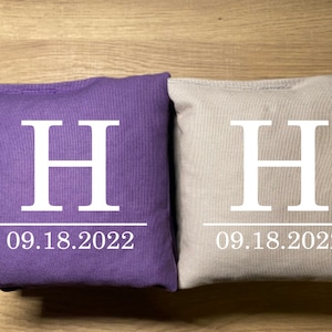 May include: Two square bean bags, one purple and one beige, are displayed. Each bag has a large white letter "H" printed on it, along with the date "09.18.2022" in white. The bags are likely for a game like cornhole.