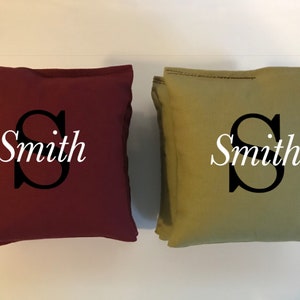 May include: Two square throw pillows, one burgundy and one olive green. Each pillow features a large black letter "S" and the name "Smith" in white script. The pillows are likely for home decor.