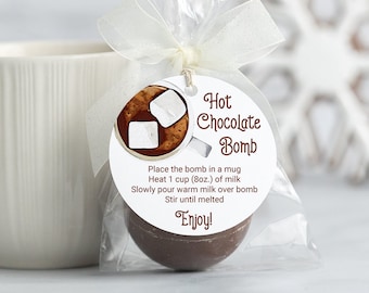 May include: A white ceramic mug stands beside a cellophane bag holding a hot chocolate bomb. The bag has a tag with "Hot Chocolate Bomb" and instructions. The background features a snowflake decoration. The tag says "Enjoy!" in white font.