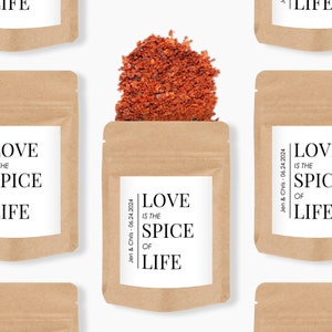May include: A brown kraft paper pouch filled with red chilli flakes. The white label features the black text "LOVE IS THE SPICE OF LIFE" and "Jen & Chris - 06.24.2024". The pouch is displayed in a repeating pattern against a plain white background.