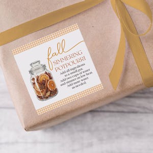Può includere: Un regalo avvolto in carta marrone con un nastro dorato. Un'etichetta quadrata presenta le parole "Fall Simmering Potpourri" e un'illustrazione di un barattolo pieno di arance secche e spezie.