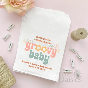 Pode incluir: Sacos de presente brancos com um design de beb&ecirc; groovy em rosa, laranja e azul com margaridas brancas. O texto diz "Obrigado por celebrar nosso beb&ecirc; groovy" e "Ch&aacute; de beb&ecirc; de Hannah & Jason 10 de janeiro de 2024".