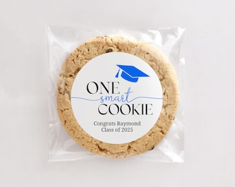 May include: A white round sticker with blue and black text that reads "ONE smart COOKIE" with a blue graduation cap graphic. The sticker is on a brown cookie in a clear plastic bag. The text below the graphic reads "Congrats Raymond Class of 2025".
