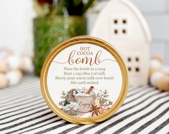 Op de afbeelding: Een ronde container met een gouden rand, met het opschrift "Hot Cocoa Bomb". Het etiket bevat instructies en een feestelijke illustratie van een mok met marshmallows, een kaneelstokje en een peperkoekmannetje. De tekst luidt "Enjoy!"
