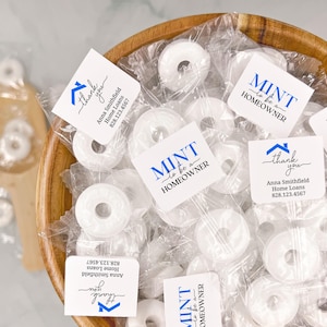 May include: A wooden bowl filled with individually wrapped white mint candies. Each candy wrapper has the text "MINT to be a HOMEOWNER" in blue and white, along with a thank you message and contact information.