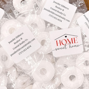 Peut inclure: Une collection de bonbons blancs en forme d'anneau emballés individuellement dans du plastique transparent. Chaque emballage comporte une étiquette carrée blanche imprimée avec le texte "HOME sweet home" et les coordonnées de Jennifer DiMarco.