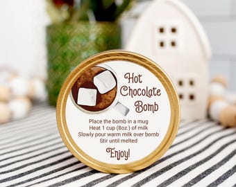 Op de afbeelding: Een ronde container met een gouden rand en een label met de tekst "Hot Chocolate Bomb". Het label toont een kop warme chocolademelk met marshmallows en instructies. De container staat op een gestreepte doek.