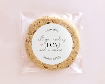 May include: A white round sticker with the text "12.20.2024 all you need is LOVE and a cookie Gretchen & Philip" with a pink heart symbol. The sticker is on a brown cookie in a clear plastic bag.