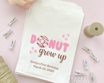 May include: White paper favor bags with a pink donut graphic and the text "Donut grow up". The bags are decorated with pink polka dots and the text "Emily's First Birthday March 10, 2023".