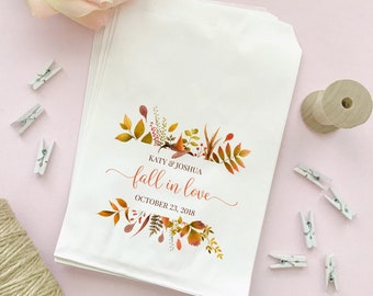 May include: White paper favor bags with a fall-themed floral design. The bags feature the text "Katy & Joshua fall in love October 23, 2018" in a script font, with autumn leaves and foliage in shades of orange, yellow, and brown.
