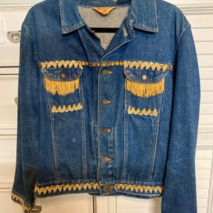 May include: A vintage denim jacket in a medium wash, embellished with gold-coloured trim and fringe. The jacket has a classic collar, button-up front, and two chest pockets. The trim and fringe provide a decorative element to the jacket.