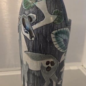 May include: A tall ceramic pitcher featuring a hand-painted scene of a deer standing among green leaves and branches. The background has a dark blue vertical line texture, while the base displays a blue cross-hatched pattern. The rim is dark blue.