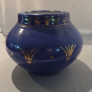 May include: A small, rounded ceramic vase with a deep blue, speckled glaze. The exterior is decorated with gold-coloured, stylised floral motifs arranged in a band around the neck and scattered across the body of the vessel.
