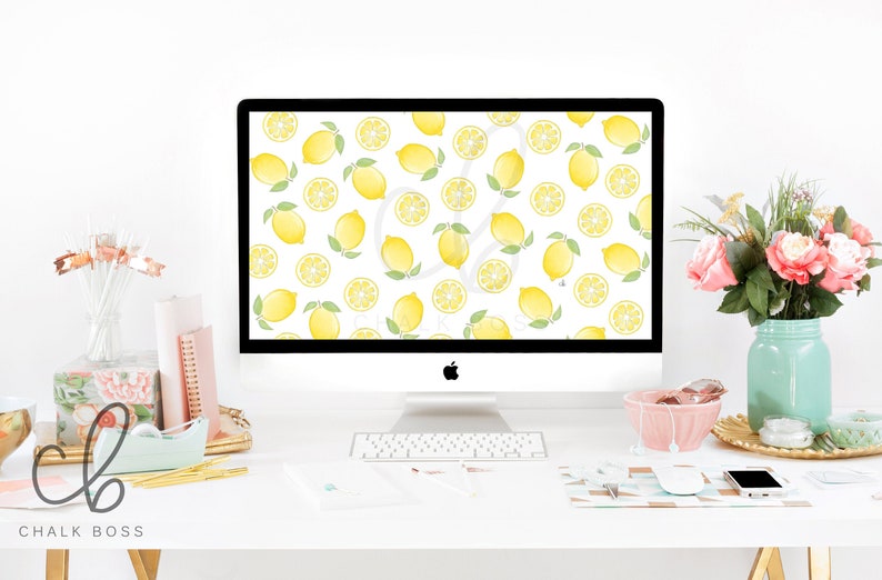 May include: A white desktop computer with a lemon pattern wallpaper. The computer is on a white desk with a keyboard, a pink bowl, a vase of pink flowers, and a gold tray with various office supplies.