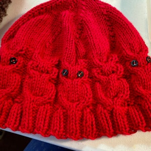 May include: A knitted red hat with an owl design. The hat features three owl faces with black button eyes. The hat is made of a thick, textured yarn and has a ruffled brim. A container of buttons is in the background.