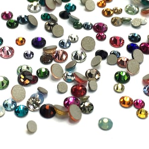 May include: A collection of colorful rhinestones in various shapes and sizes, including round and oval. The gems are in shades of pink, green, blue, purple, and gold, with a flat, silver backing. The rhinestones are scattered on a white surface.