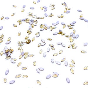 Può includere: Una collezione di piccoli strass a forma di marquise sparsi nell'inquadratura. Ogni pezzo presenta una superficie superiore gialla traslucida sfaccettata e un retro piatto di colore argento opaco, creando un effetto scintillante.