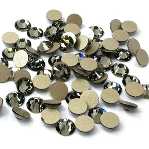 May include: A pile of black diamond-shaped rhinestones with a silver backing. The rhinestones are scattered on a white surface.