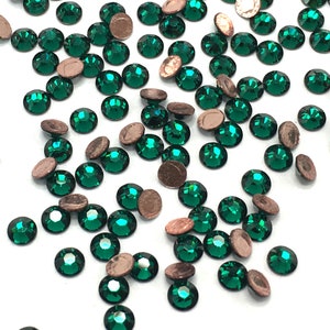 Puede incluir: Una dispersión de piedras de imitación de color verde esmeralda con engastes de cobre. Las piedras de imitación están dispersas sobre un fondo blanco.