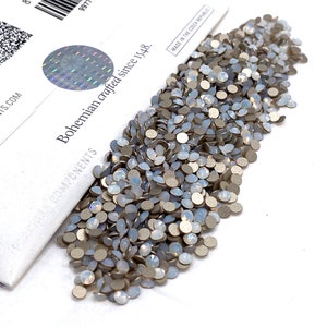 May include: A large pile of white opal rhinestones with a silver backing. The rhinestones are scattered on a white surface. The rhinestones are packaged in a white box with the text "Bohemian crafted since 1548." and "Made in the Czech Republic."