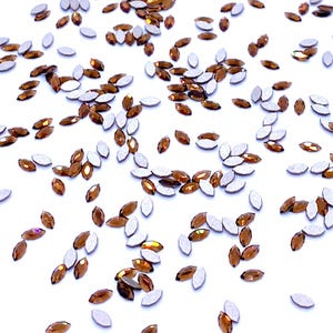 Può includere: Una collezione di numerosi piccoli cristalli color ambra a forma di marquise sparsi su una superficie bianca uniforme. Ogni cristallo presenta una parte superiore sfaccettata marrone traslucida e un retro piatto di colore argento opaco.