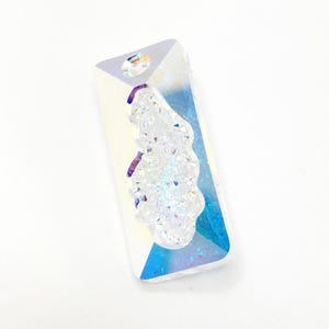 Puede incluir: Colgante rectangular de cristal con un agujero en la parte superior. El colgante tiene un acabado transparente e iridiscente con un grupo de material blanco y brillante en el centro. La esquina inferior derecha tiene un tono azul.