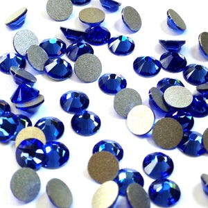 May include: A collection of scattered, faceted, sapphire blue rhinestones with silver backing. The gemstones are oval and round, reflecting light. The image is a close-up, showcasing the sparkle and detail of the crystals, ideal for craft projects.
