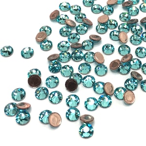 May include: A scattering of turquoise blue rhinestones with silver backing. The rhinestones are scattered on a white surface.