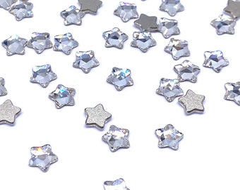 May include: A scattering of clear star-shaped rhinestones on a white background. The rhinestones have a silver backing.