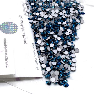 Puede incluir: Un montón de pedrería autoadhesiva azul oscuro con respaldo blanco. La pedrería está esparcida sobre una superficie blanca. El embalaje de la pedrería es visible en el fondo. El embalaje es blanco con texto negro que dice "PRECiosa COMPONENTS" y "Bohemian crafted since 1948. MADE IN THE CZECH REPUBLIC."