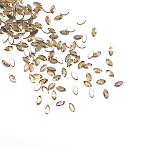 May include: A scattering of small, iridescent, marquise-shaped rhinestones in a light brown colour. The rhinestones are scattered on a white background.