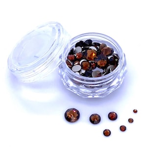 May include: A clear, faceted plastic container with an open lid, filled with various sizes of brown and silver rhinestones. Several rhinestones are scattered around the container. The rhinestones have a faceted, reflective surface, and are arranged on a white background.