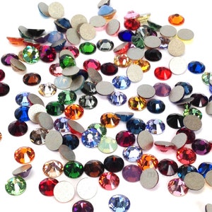 Pode incluir: Conjunto de strass coloridos em vários formatos e tamanhos, incluindo redondos, quadrados e ovais. Os strass estão espalhados sobre um fundo branco.