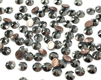 May include: A collection of dark gray, faceted rhinestones with a metallic sheen. Some rhinestones have a copper-colored backing, scattered on a white surface. These embellishments are suitable for various craft projects.