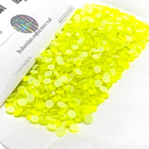 Puede incluir: Primer plano de una gran cantidad de pequeños diamantes de imitación redondos, planos y de color amarillo brillante. Los diamantes de imitación están dispuestos en una pila sobre una superficie blanca. Los diamantes de imitación están hechos en la República Checa.