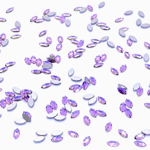 Può includere: Una collezione di piccoli strass viola a forma di marquise sparsi nell'inquadratura. Ogni singolo cristallo presenta una superficie superiore sfaccettata in una tonalità viola vibrante e un retro metallico color argento piatto per il fissaggio.