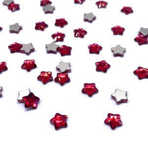 May include: Red star-shaped rhinestones scattered on a white background. The rhinestones have a faceted design and a silver backing.