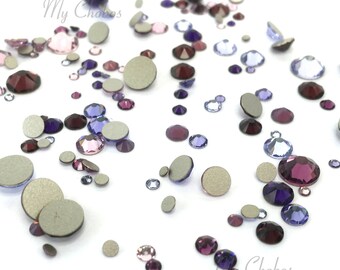 May include: Assortment of small, round, iridescent rhinestones in shades of purple, pink, and blue. The rhinestones are scattered on a white surface.